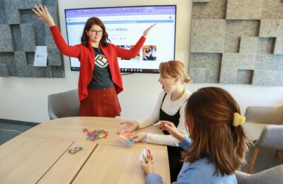 Three women engage in a lively team building card game in a modern office setting.