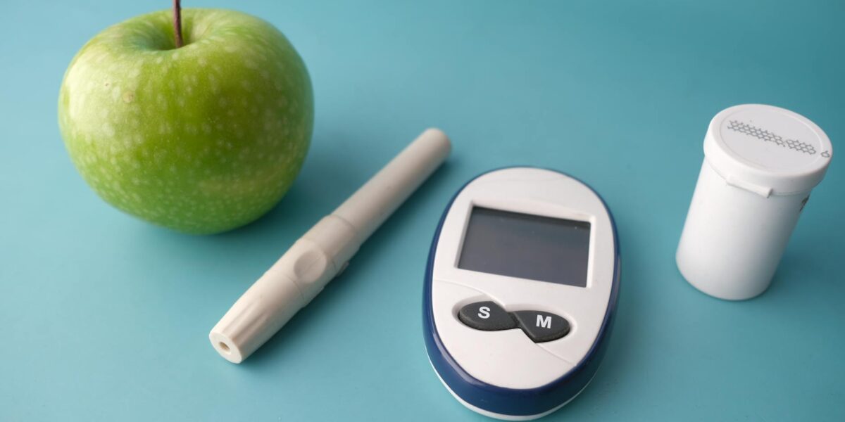 Flat lay of a glucose monitor, apple, and insulin pen on a pastel background.