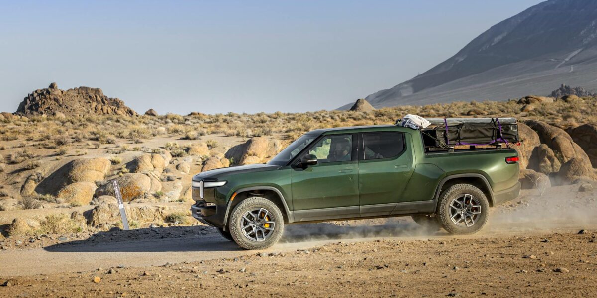 Green electric pickup truck on a dusty trail in a rocky desert landscape.