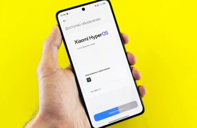 Hand holding smartphone showing Xiaomi HyperOS update screen on yellow background.
