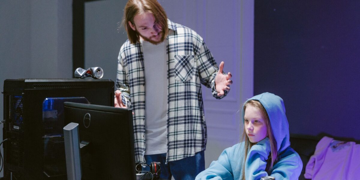A father scolds his daughter playing video games on a computer, creating a tense moment.