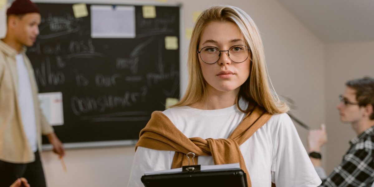 A young woman wearing glasses stands in a classroom holding a clipboard.