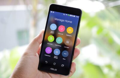 A hand holding a smartphone displaying home management apps on the screen.