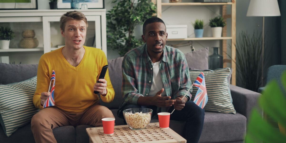 Two men sitting on a sofa, watching TV with popcorn and flags, enjoying a sports event indoors.