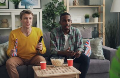Two men sitting on a sofa, watching TV with popcorn and flags, enjoying a sports event indoors.