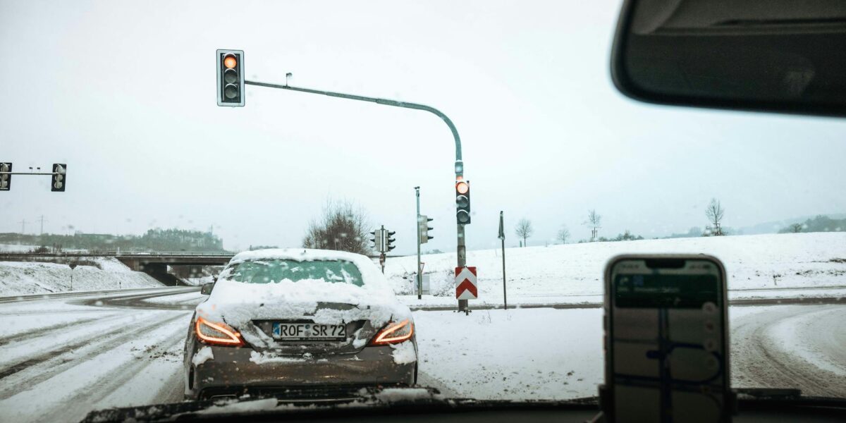 A car waits at a traffic light during snowfall on a snowy road, depicting winter driving conditions.