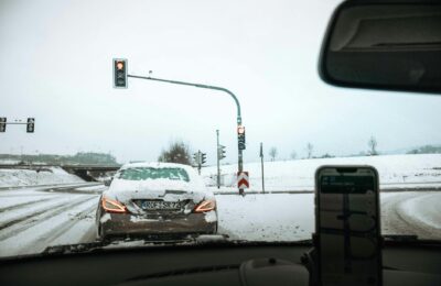 A car waits at a traffic light during snowfall on a snowy road, depicting winter driving conditions.