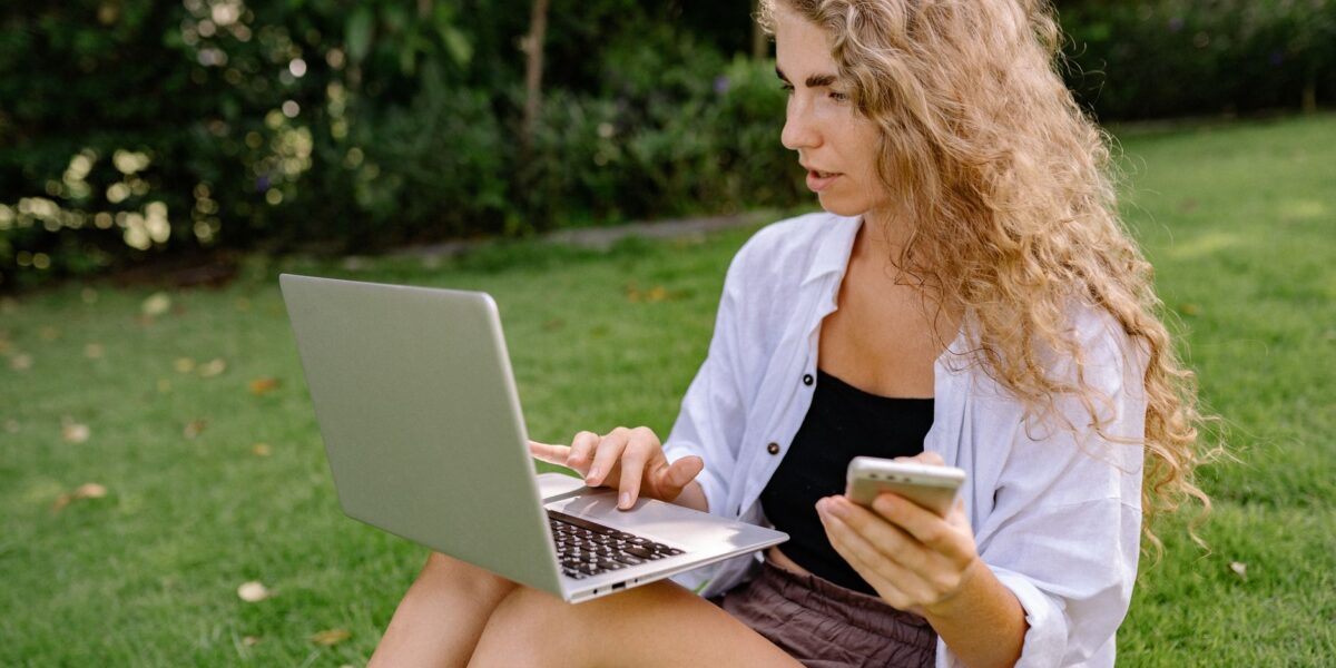 Pensive female remote employee with cellphone working on portable computer while sitting on grass on meadow near trees in daylight