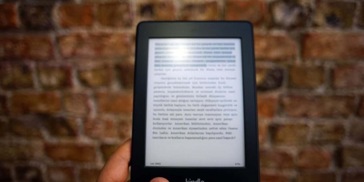 Close-up of a Kindle e-reader held by a person, showcasing a digital reading device against a textured brick wall backdrop.