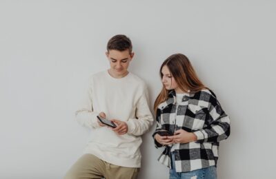 Teenage boy and girl using smartphones while leaning against a white wall indoors.