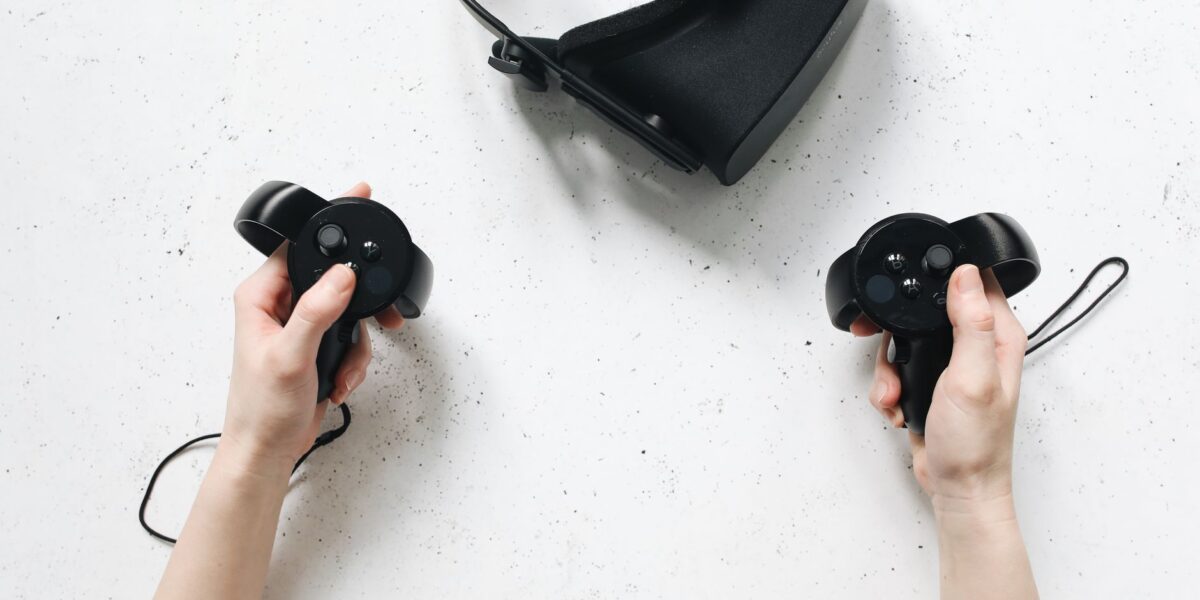 Two hands hold virtual reality controllers above a VR headset, showcasing modern technology.
