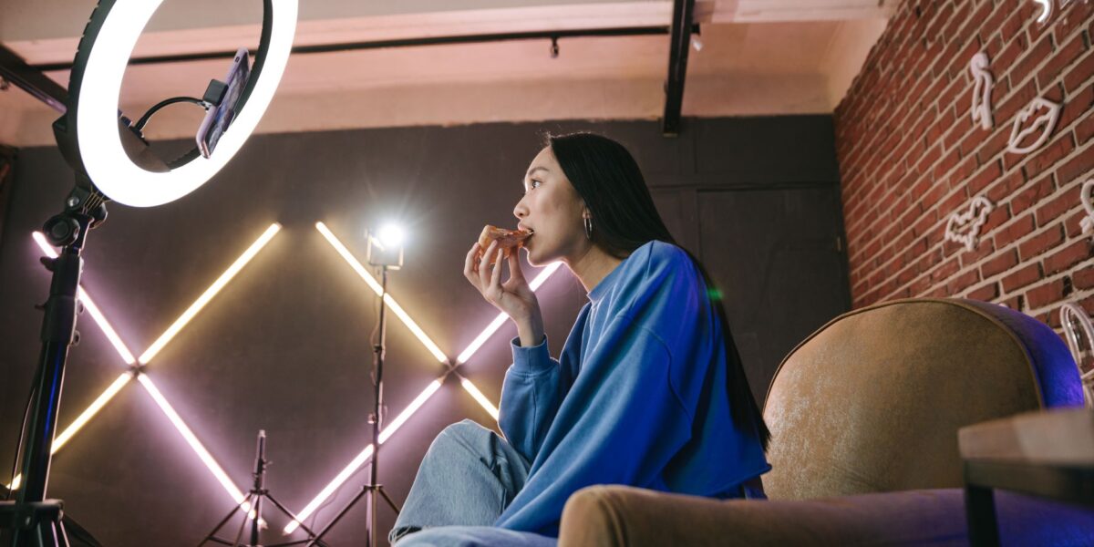 Asian woman content creator eating pizza while recording a video indoors with ring light.