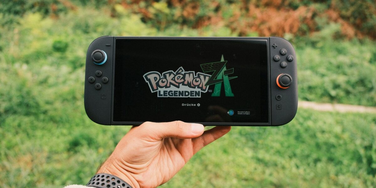 A hand holding a handheld gaming console displaying the Pokémon Legends game screen outdoors.