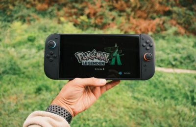 A hand holding a handheld gaming console displaying the Pokémon Legends game screen outdoors.