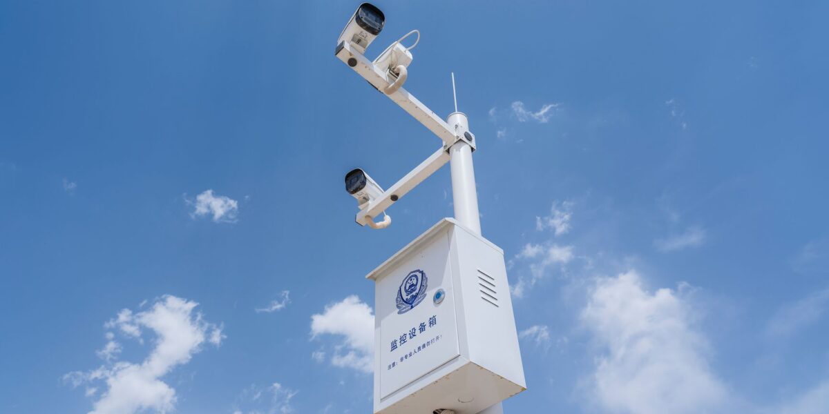 White security cameras mounted on a pole against a bright blue sky with fluffy clouds.