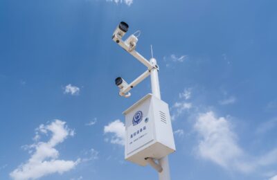 White security cameras mounted on a pole against a bright blue sky with fluffy clouds.