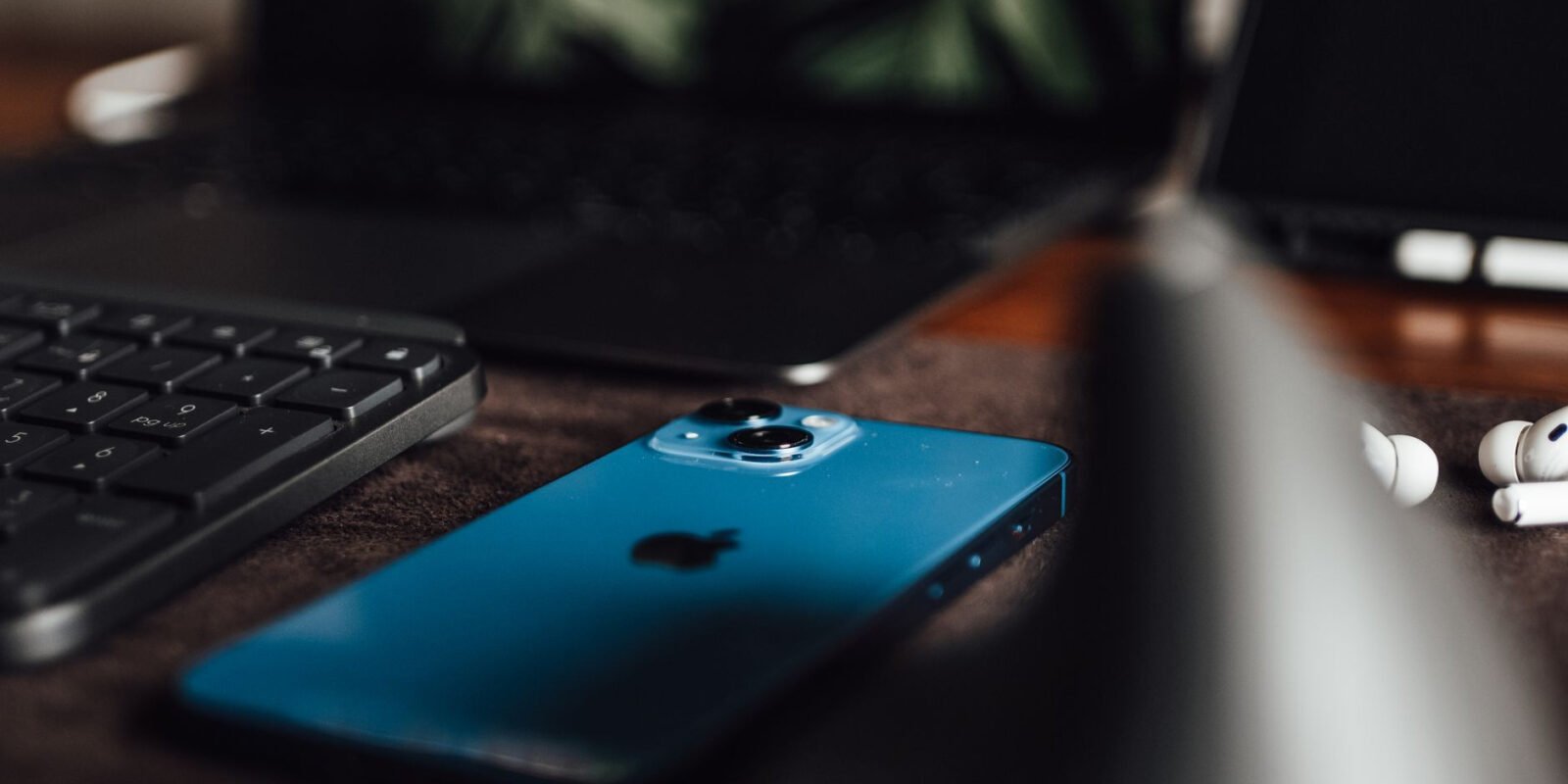 Close-up of an Apple iPhone, keyboard, laptop, and AirPods on a desk.