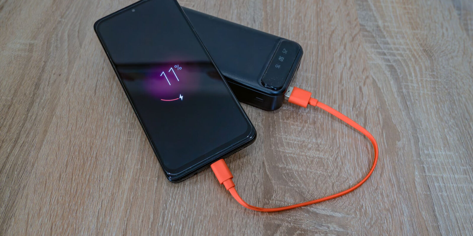 A smartphone charging from a power bank with an orange cable on a wooden table.