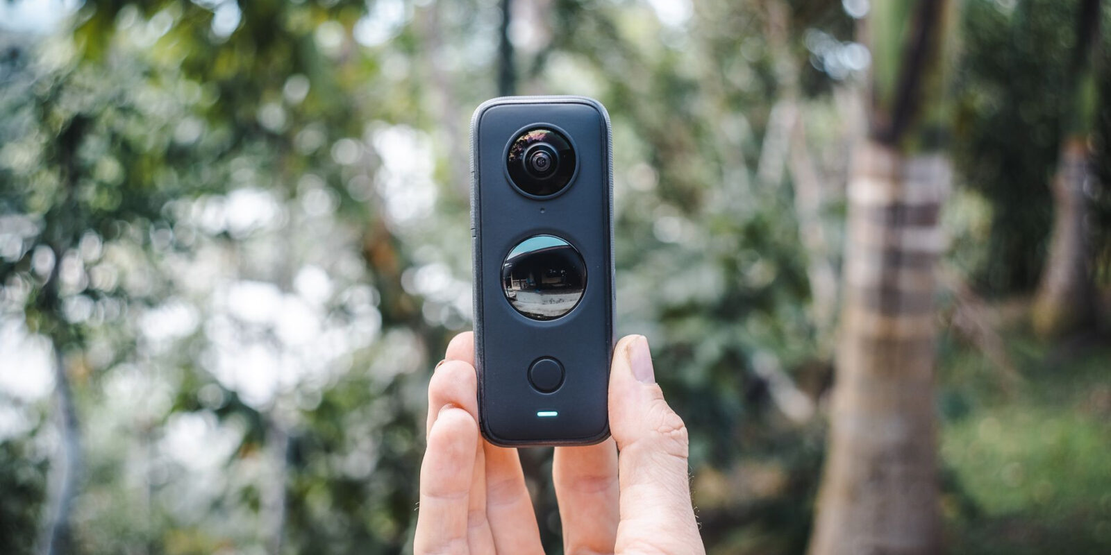 Close-up of a hand holding a compact 360 camera outdoors with greenery.