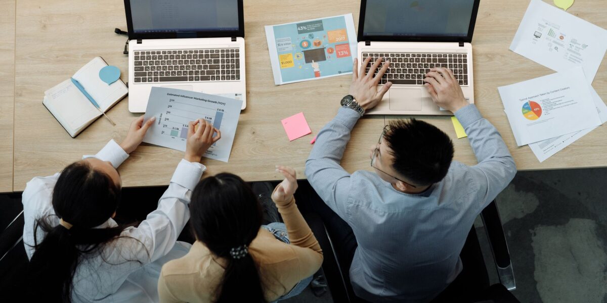 Three colleagues working on laptops and documents at a desk, collaborating on business projects.