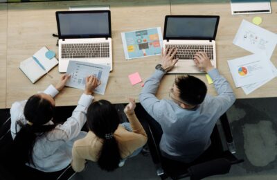 Three colleagues working on laptops and documents at a desk, collaborating on business projects.