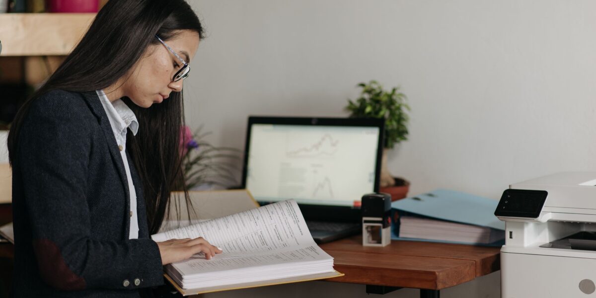 Woman reading documents at office desk with computer and printer.