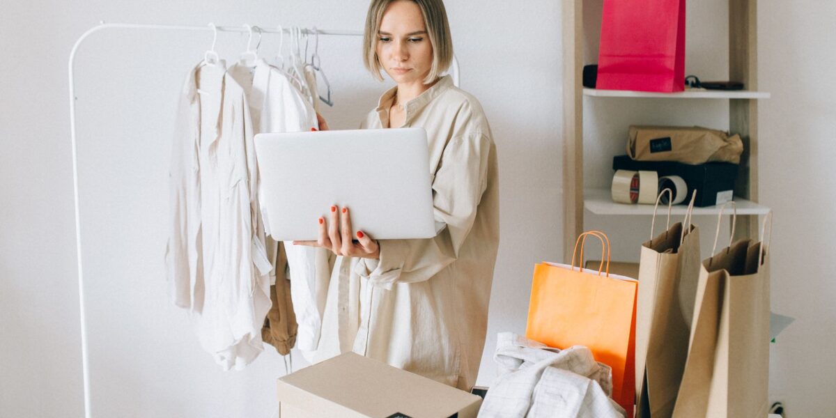 Woman using laptop for online shopping with clothes and bags around her, indoors.