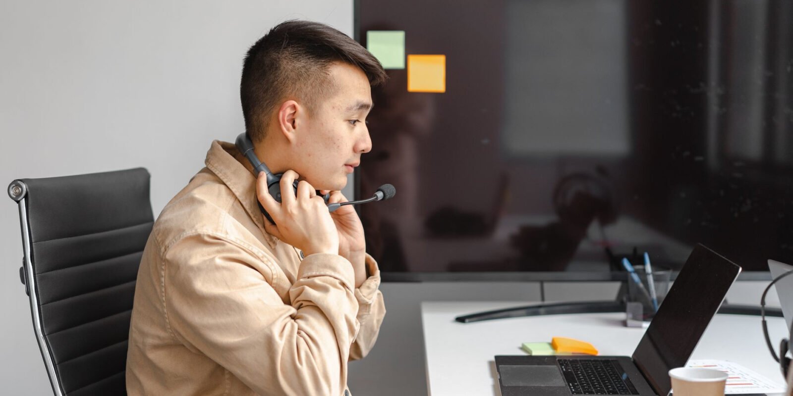 Man working with laptop in office setting, wearing a headset for an online meeting.